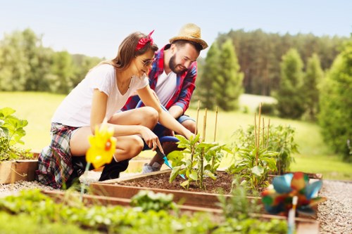 In a well-maintained outdoor garden, a man and a woman are engaged in planting or tending to young vegetable or flower plants within a raised bed area constructed of wooden planks. The raised bed contains dark, fertile soil with emerging green shoots visible. The garden margin features a gravel pathway, and beyond the planting area, there are lush green lawns and a backdrop of mature trees with dense foliage under a bright, clear sky, indicating a sunny day. The garden appears spacious, with natural light highlighting various shades of green and brown, creating a vibrant and inviting outdoor environment suitable for gardening or landscaping services offered by Garden Maintenance Crystal Palace. The scene captures a moment of outdoor activity in a serene landscaped yard, emphasizing the natural textures of soil, plants, and gravel, with a natural and professional tone suitable for a gardening service website in the Crystal Palace area, London postcode SE19 or nearby.
