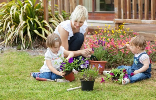 A woman with light blonde hair and two young girls with light brown hair are engaged in gardening activities in a well-maintained backyard garden in Crystal Palace. The woman is kneeling on the grass, smiling as she helps the children plant and tend to a variety of potted flowering plants, including purple and pink blooms. The garden features a lush, evenly cut green lawn with a neat border of ornamental grasses and shrubs, which are visible in the background. Behind the garden, there is a wooden deck with steps leading down to the grassy area, and a building with large glass doors, possibly a conservatory or extension, can be seen. The outdoor space is bright and lively, suggesting a clear day with natural sunlight illuminating the scene, making it suitable for professional gardening and outdoor maintenance services provided by Garden Maintenance Crystal Palace. The scene captures a moment of outdoor family gardening activity, emphasizing natural textures, vibrant flower colours, and a clean, structured garden layout in an urban residential environment in South London postcode starting with SE, aligning with local gardening services.