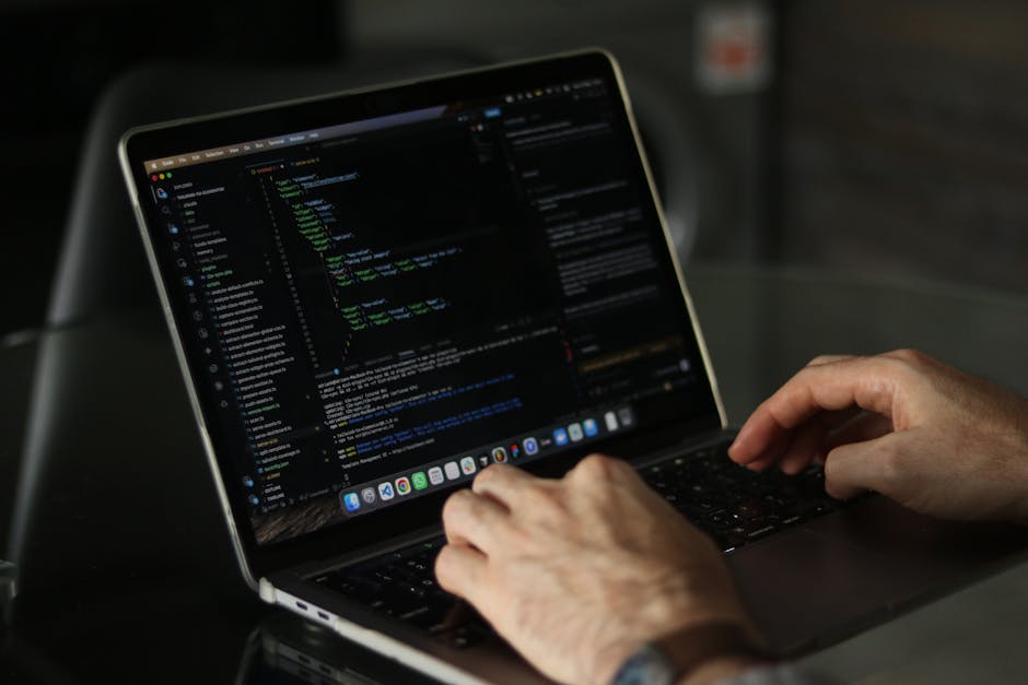 A close-up of a person's hands typing on a laptop keyboard, displaying lines of code on the screen. The laptop is placed on a dark surface in a dimly lit environment, with the screen showing a dark-th