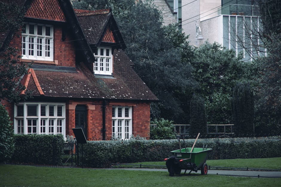 A Victorian-style brick house with a steep, tiled roof and white-framed sash windows, surrounded by a well-maintained garden. The garden features neatly trimmed hedges and a lush green lawn with textu