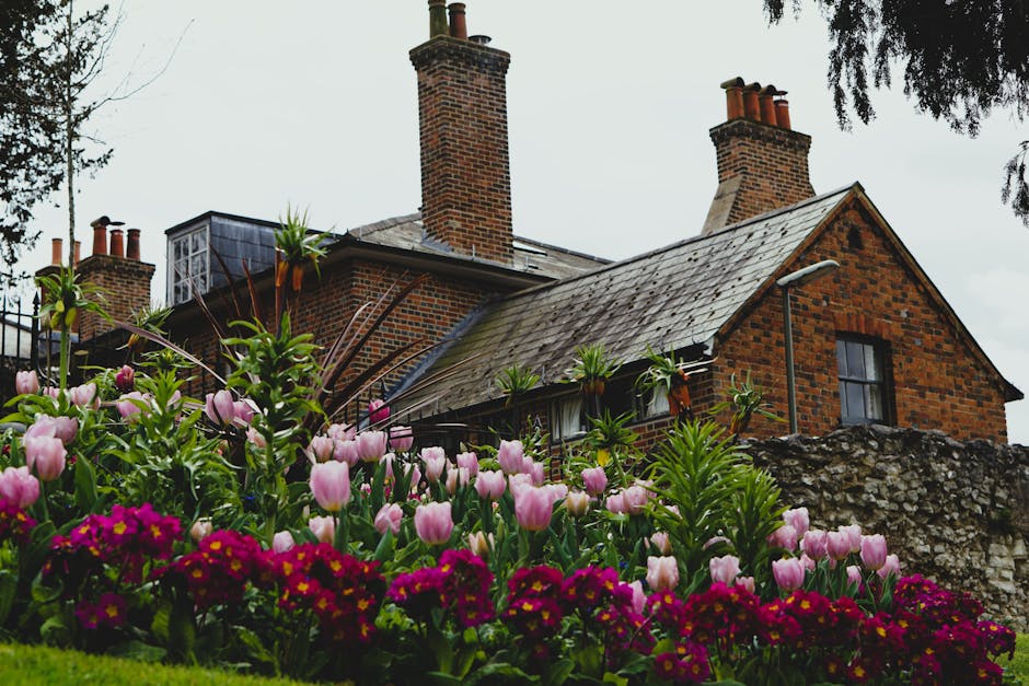 A Victorian-style brick house with a steep, tiled roof and white-framed sash windows, surrounded by a well-maintained garden. The garden features neatly trimmed hedges and a lush green lawn with textu