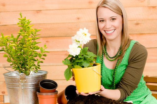 The image depicts a woman with blonde hair and a bright smile, sitting outdoors in a garden setting with a wooden fence background. She is wearing a green polka-dot top and layered necklaces, holding a yellow flower pot with a small, vibrant green plant or flowering shrub. To her left, there is a metallic bucket containing another green shrub, and nearby are empty plant pots of various sizes, including black and terracotta-colored ones. The garden area features a well-maintained lawn with lush green grass, bordered by flower beds or shrubbery. The scene appears to be under natural daylight with a clear, mild weather condition. This setting highlights a typical garden space suitable for landscaping, planting, and outdoor maintenance, aligning with services offered by Garden Maintenance Crystal Palace, and subtly emphasizing gardening tasks such as potting and planting in a pleasant outdoor environment near Crystal Palace.